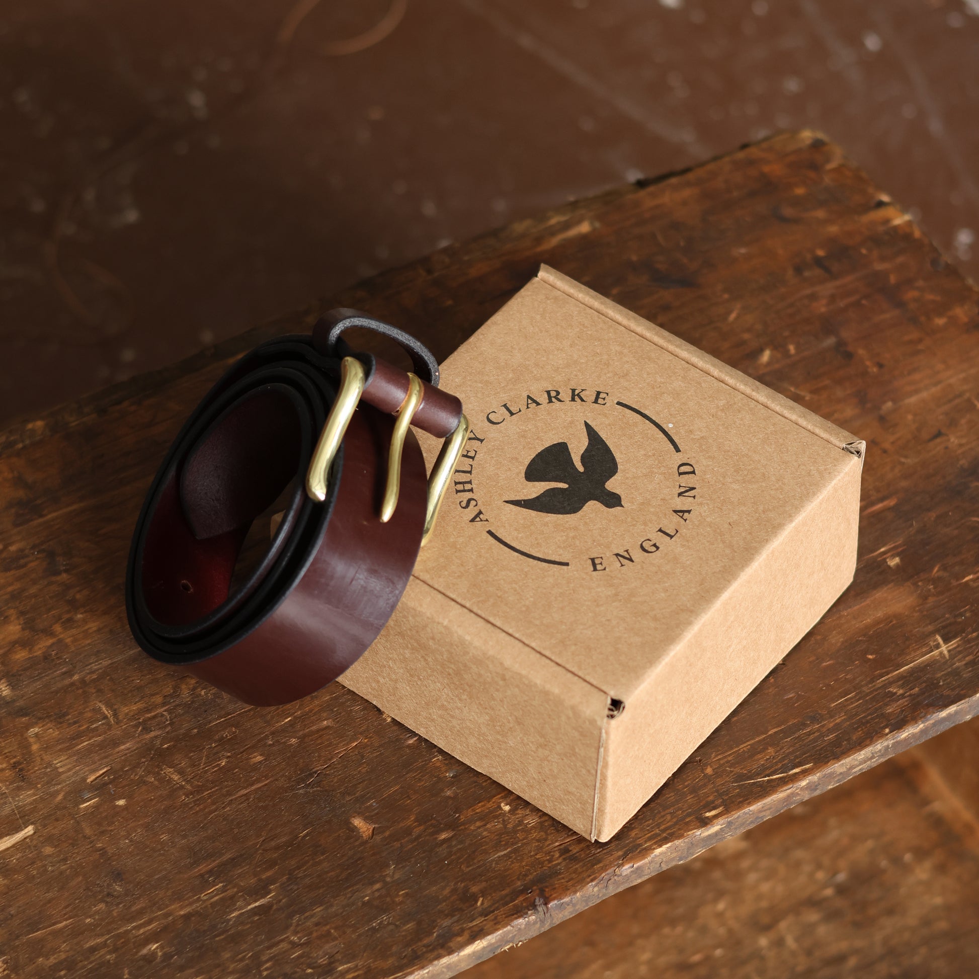 Brown leather belt with brass buckle on a cardboard box labeled 'Ashley Clarke England' on a wooden surface.